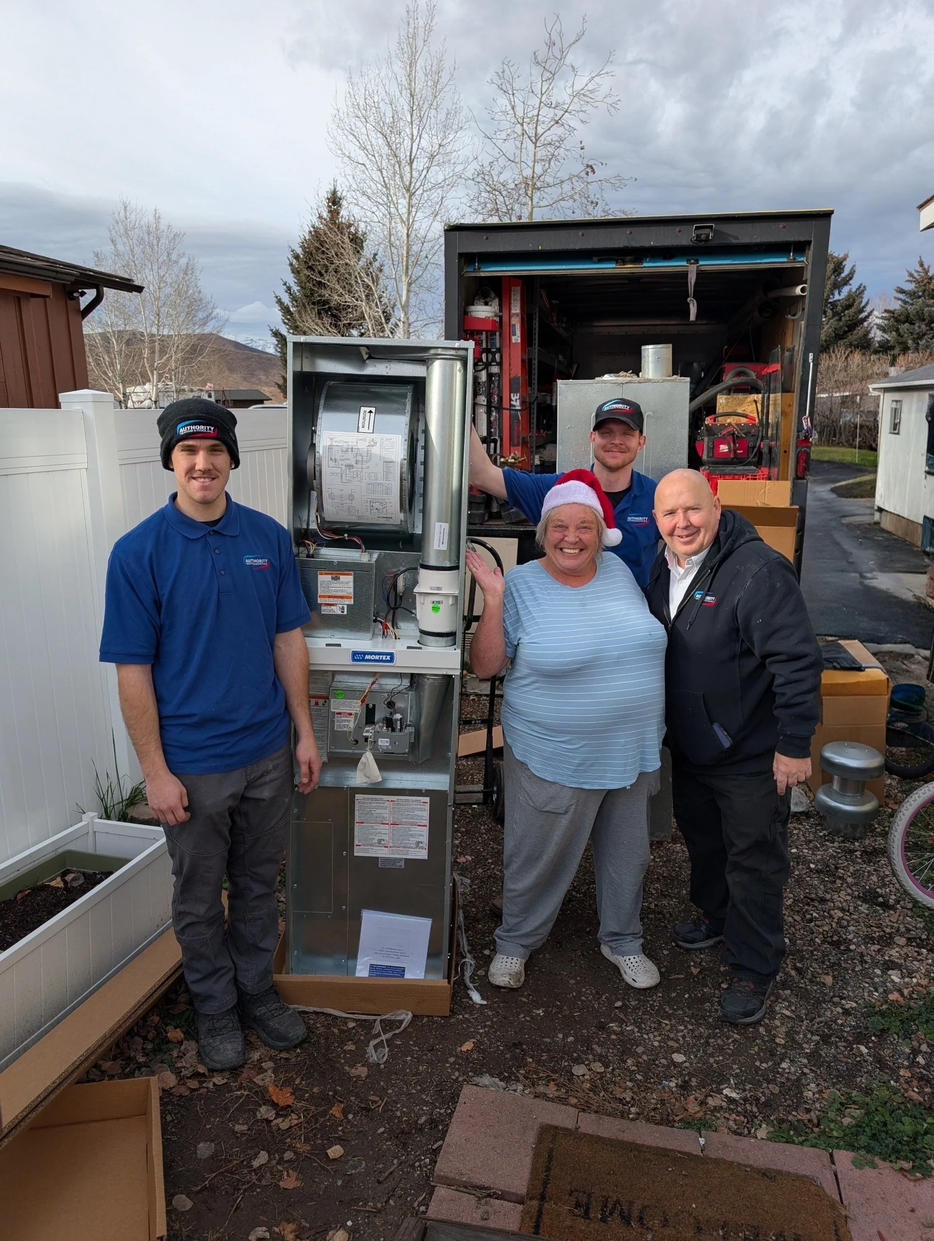 Authority Heating & Cooling technicians and a smiling homeowner in a Santa hat beside her new furnace